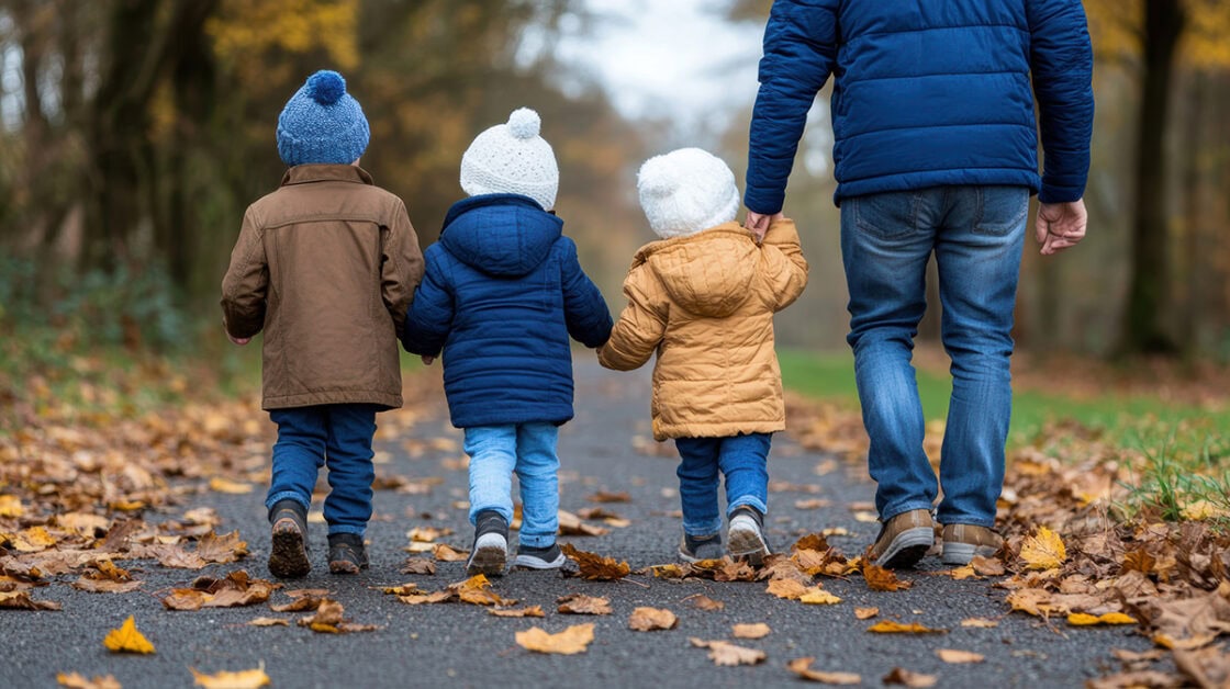 un padre con tres niños pequeños de la mano caminando por un paseo con hojas de otoño en el suelo. Se les ve de espaldas con abrigo y gorro